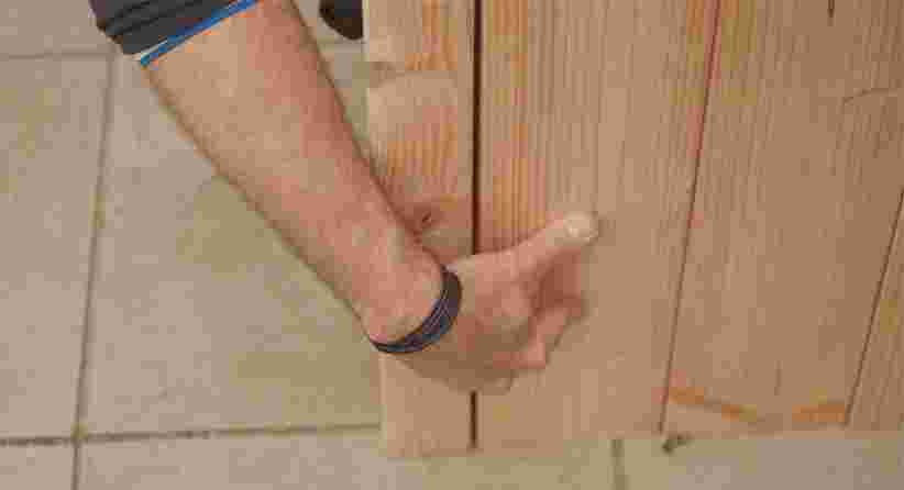 Een hand met een armband plaatst of controleert een houten plank tegen een wand op een tegelvloer.