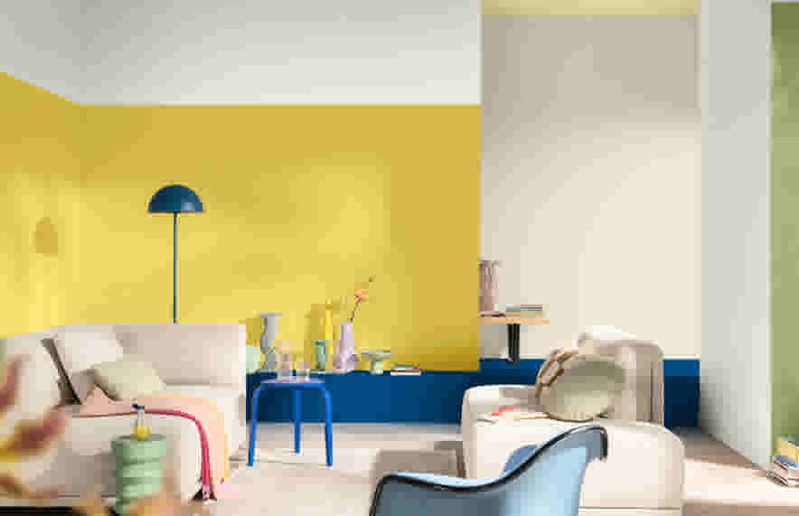 Salon moderne avec mur jaune vif, canapé beige, lampe bleue et table d'appoint bleue, créant une ambiance colorée et lumineuse.