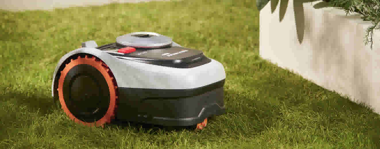 Robotmaaier met oranje wielen rijdt over groen gazon langs een witte plantenbak met kruiden.