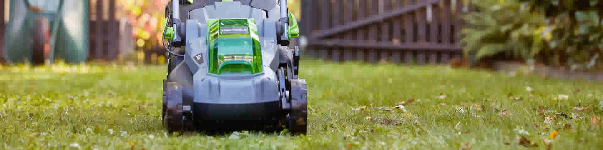 Grijze en groene elektrische grasmaaier met LUX-TOOLS logo in actie op een zonnig gazon naast een houten schutting.