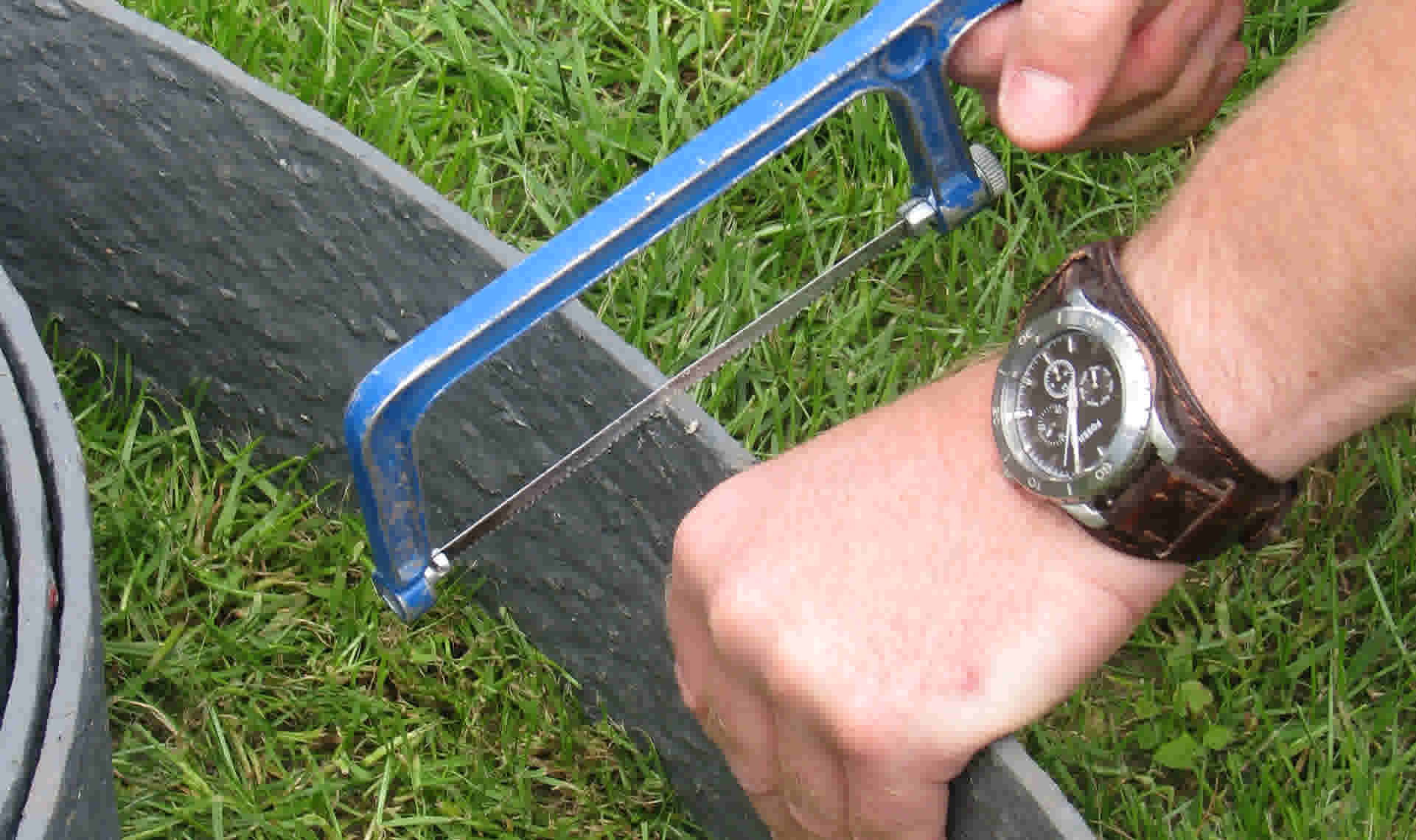 Hand met horloge gebruikt een blauwe ijzerzaag om een tuinafboording op gras te zagen.