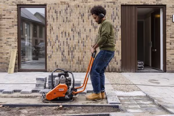 Persoon met koptelefoon bedient een gehuurde oranje trilplaat van RentPartner Boels voor bestrating voor een huis.