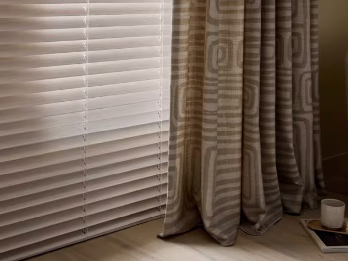 Witte jaloezieën naast beige gordijnen met geometrisch patroon, met een kopje op een dienblad op de vloer.