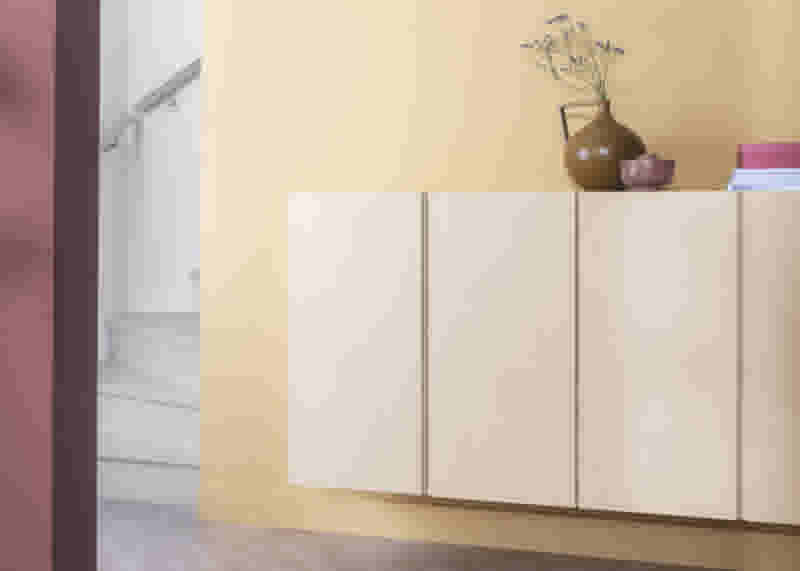 Licht houten dressoir tegen gele muur met bruine vaas en roze schaal. Op de achtergrond een witte trap.