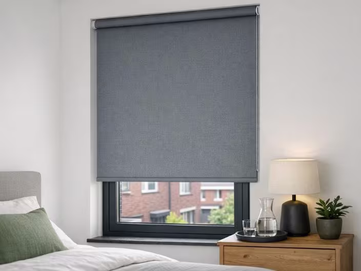 Grijze rolgordijn half neergelaten voor een raam in een slaapkamer, met een nachtkastje met lamp en plant ernaast.