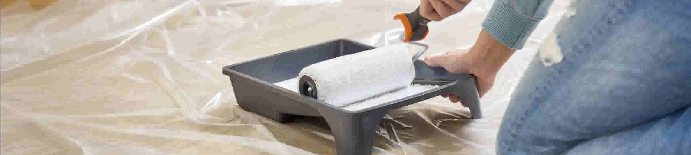 Personne préparant un rouleau à peinture blanc dans un bac gris sur une surface protégée par une bâche en plastique.
