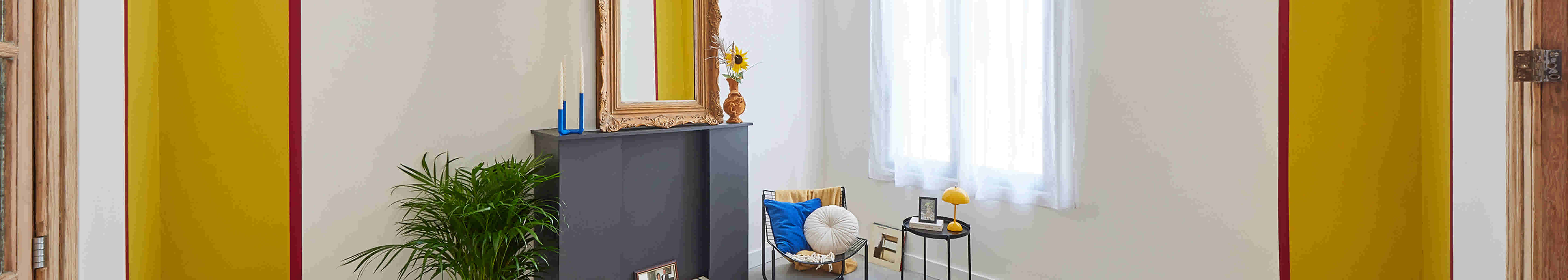 Intérieur lumineux avec murs jaunes, cheminée grise, miroir doré, plante verte et fauteuil garni de coussins bleus près d'une fenêtre.