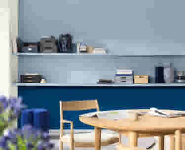 Moderne eetkamer met houten tafel, blauwe muur en witte planken met opbergdozen en decoratieve items.