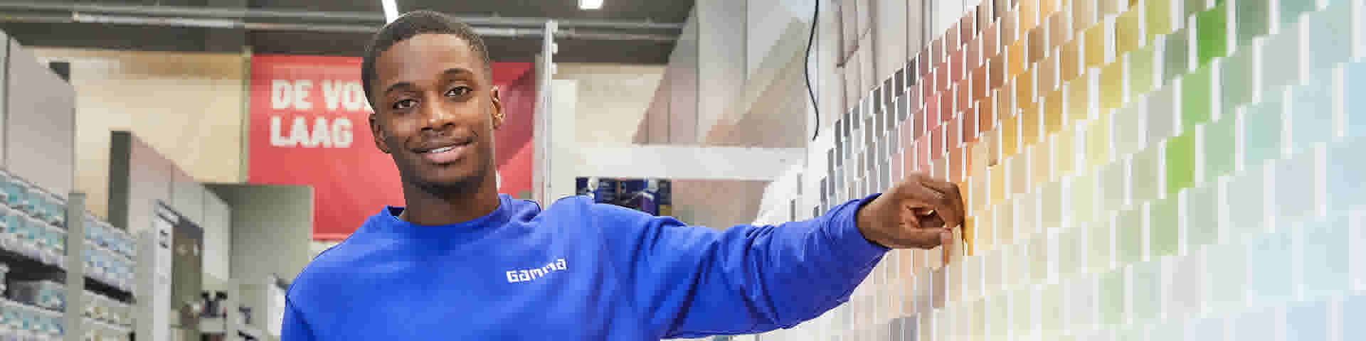 Gamma medewerker in blauw shirt wijst glimlachend naar een kleurenwaaier met verfstalen in een bouwmarkt.
