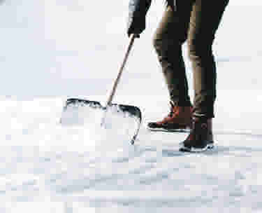Persoon in grijze jas schept sneeuw weg met een zwarte sneeuwschuiver voor een wit huis met grote ramen.