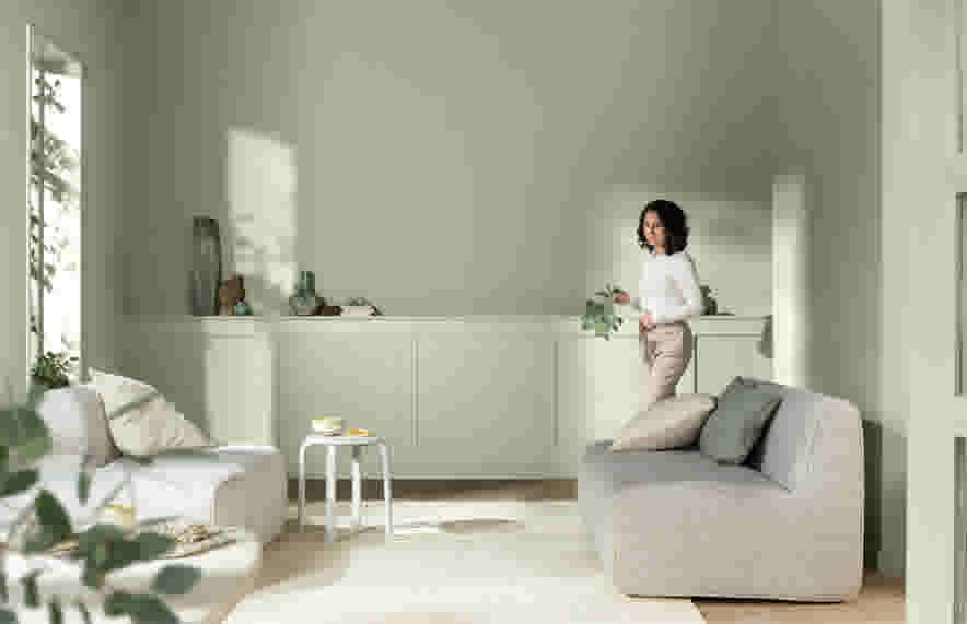 Vrouw in witte blouse staat in lichte, moderne woonkamer met beige banken, witte kast en natuurlijk licht door raam.
