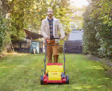 Man maait het gazon met een rode grasmaaier in een zonnige achtertuin omringd door groene struiken.