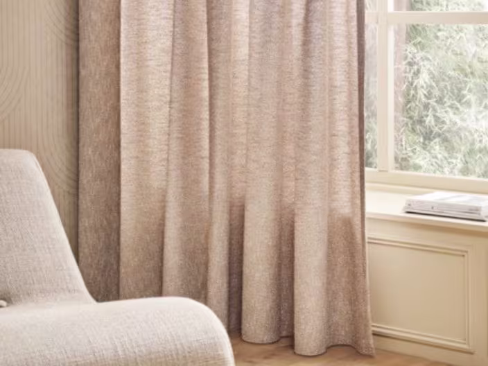 Beige gordijn naast een raam met natuurlijk licht, een lichte stoel links en een vensterbank rechts in een rustige woonkamer.
