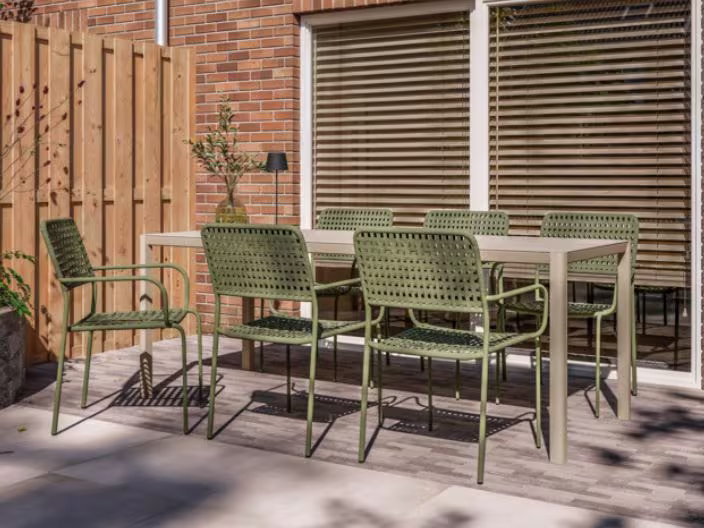Gezellig terras met groene tuinstoelen en tafel tegen bakstenen muur, houten schutting en zonwering voor het raam.