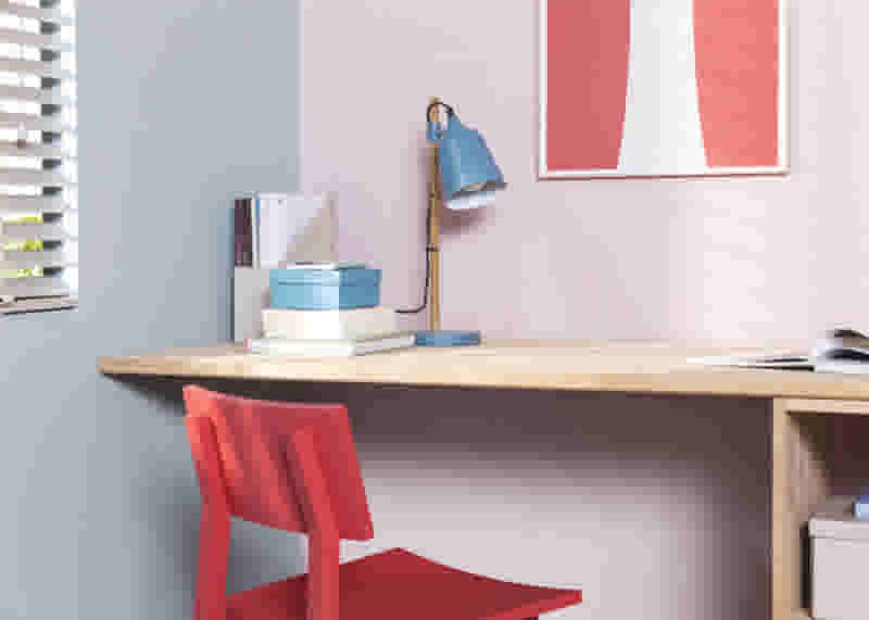 Houten bureau met blauwe lamp, boeken en rode stoel. Roze muur met kunstwerk. Lichte, moderne werkruimte.