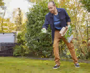 Homme en veste bleue et pantalon marron épandant des produits sur sa pelouse dans un jardin entouré d'arbustes verts.
