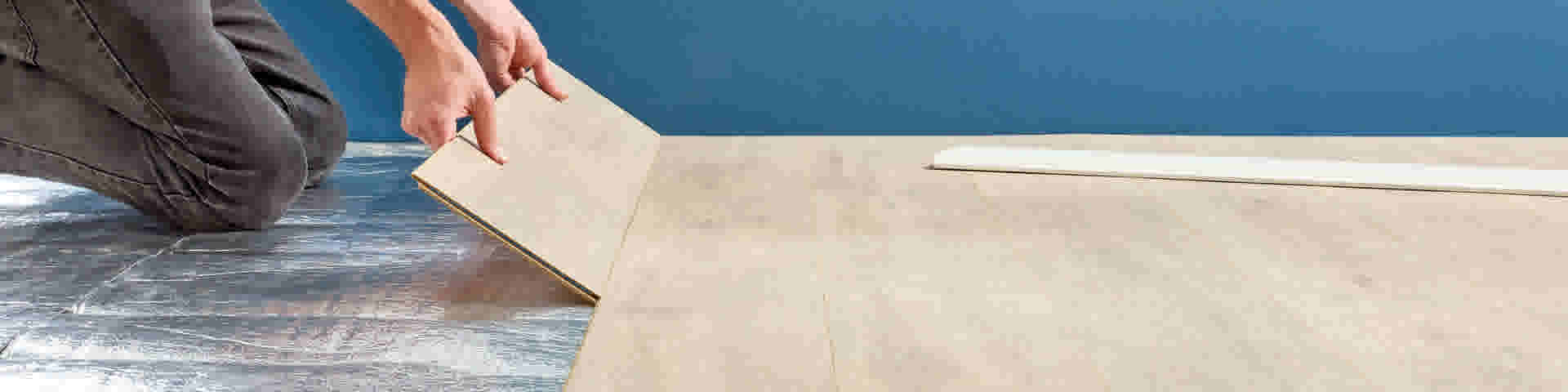 Personne en jean gris installant un plancher stratifié beige sur une sous-couche argentée contre un mur bleu.