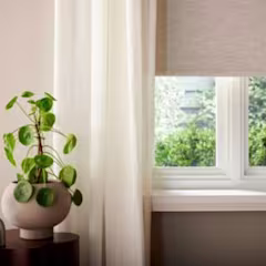 Store enrouleur beige à mi-hauteur, rideau blanc léger, plante verte en pot rond beige sur table sombre près d'une fenêtre lumineuse.