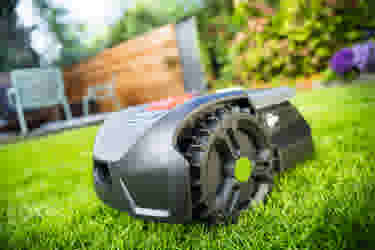 Robotmaaier op groen gazon in een achtertuin met houten schutting en tuinstoelen op de achtergrond.