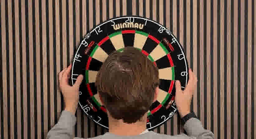Persoon hangt een Winmau dartbord op aan een gestreepte muur, beide handen houden het bord vast.