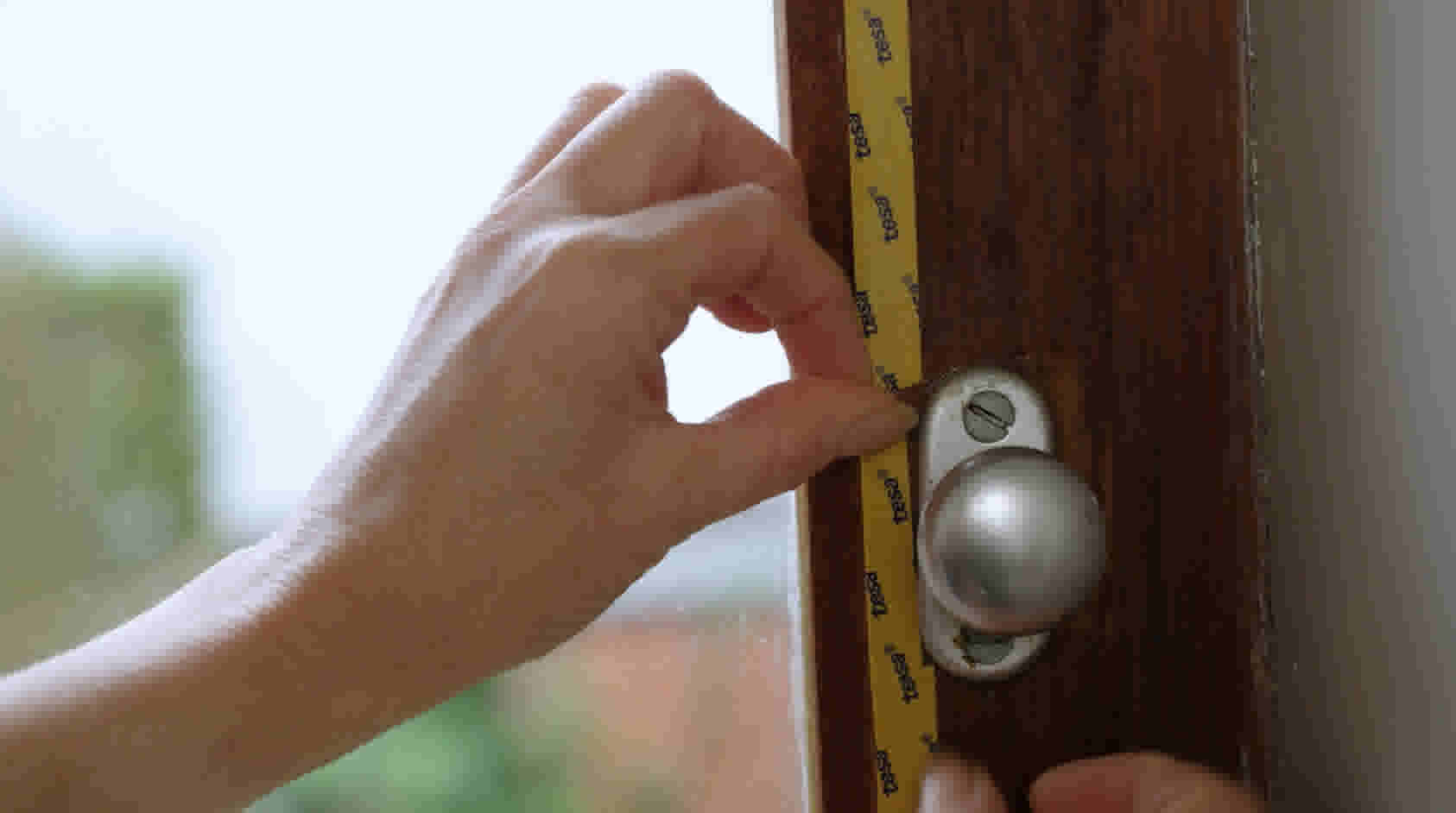 Hand plaatst gele tesa-tape langs een houten raamkozijn met metalen draaiknop voor betere isolatie.