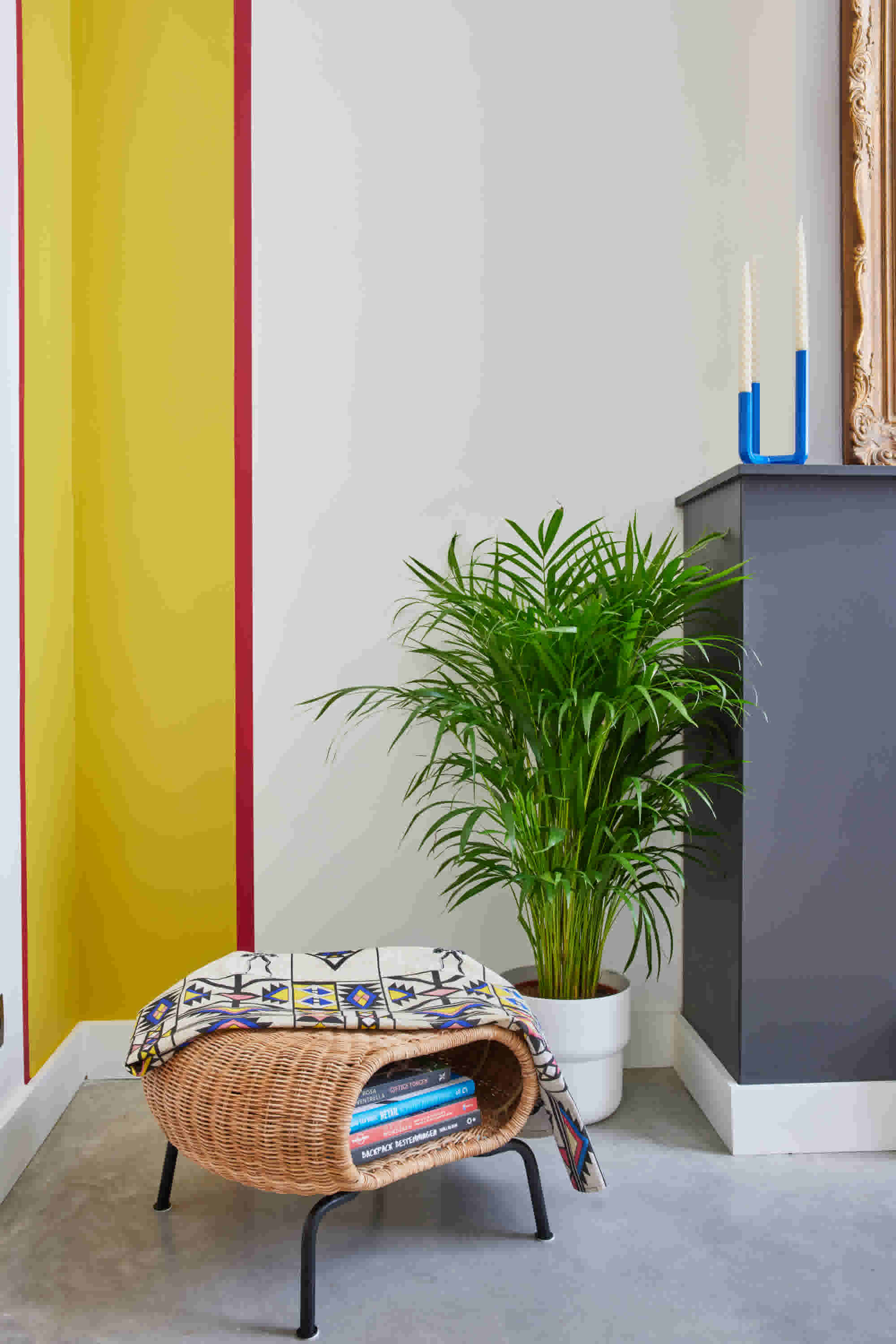 Moderne woonhoek met rieten bijzettafel, boeken, kleurrijke deken, groene kamerplant en blauwe kandelaars op grijs meubel.