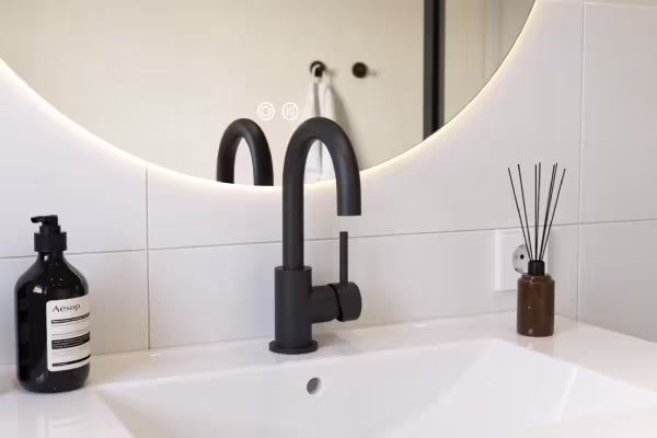 Moderne badkamer met zwarte kraan, witte wastafel, ronde verlichte spiegel, zeeppompje en geurstokjes in bruine fles.