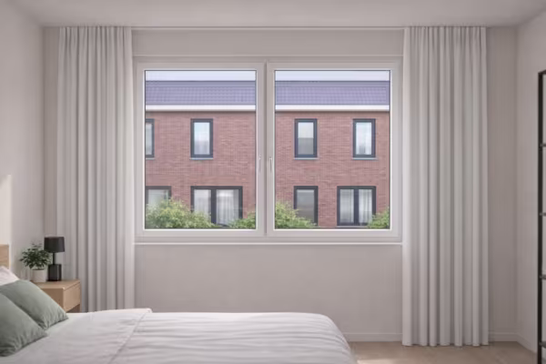 Lichte slaapkamer met groot raam dat uitkijkt op bakstenen huizen, witte gordijnen en een comfortabel bed met groene kussens.