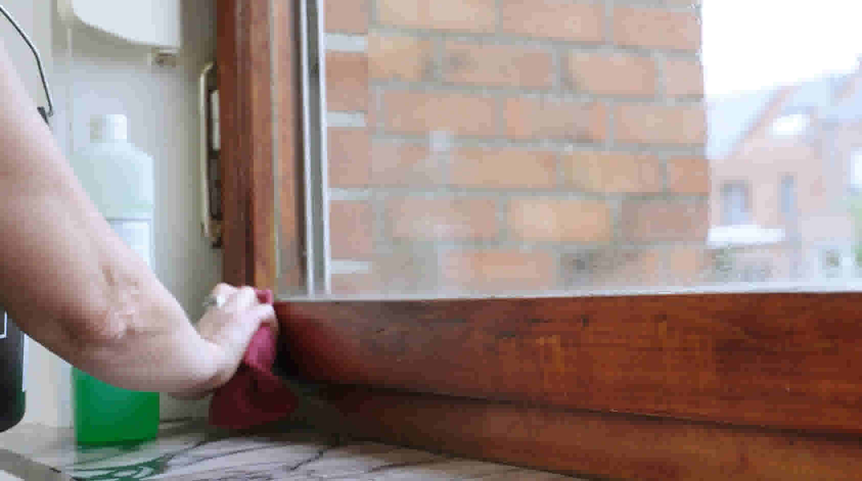 Hand poetst houten vensterbank met rode doek naast reinigingsflessen, met bakstenen muur zichtbaar door het raam.