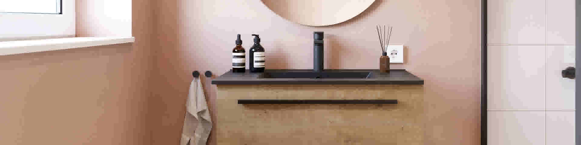 Salle de bain moderne avec murs roses, meuble en bois, lavabo noir, miroir rond et flacons élégants de produits cosmétiques.