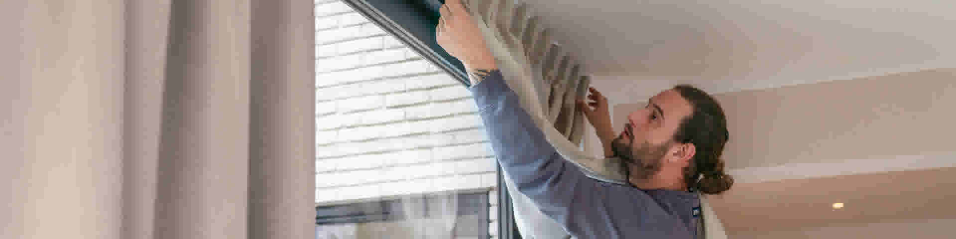 Persoon in blauw shirt hangt beige gordijnen op aan een zwarte gordijnrail bij een raam met witte jaloezieën.