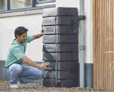 Homme en t-shirt vert accroupi installant un récupérateur d'eau de pluie gris contre le mur d'une maison.