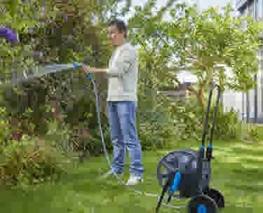 Persoon in lichte kleding geeft planten water met een tuinslang in een groene tuin, met een slanghaspel op wielen ernaast.