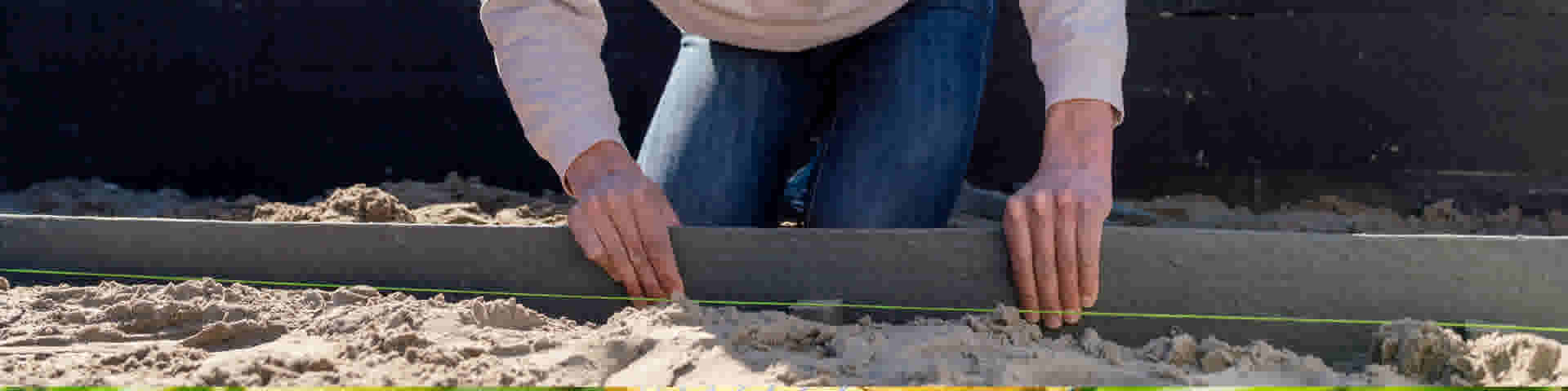 Personne en jean et pull blanc nivelant une Bordure Ecolat frais avec une règle sur un chantier de construction.