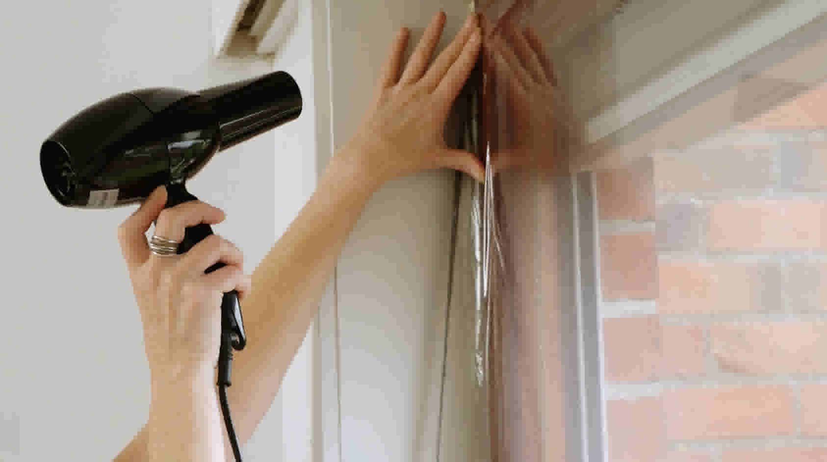 Persoon installeert isolatiefolie op raam met föhn om luchtbellen te verwijderen en folie strak te krijgen.