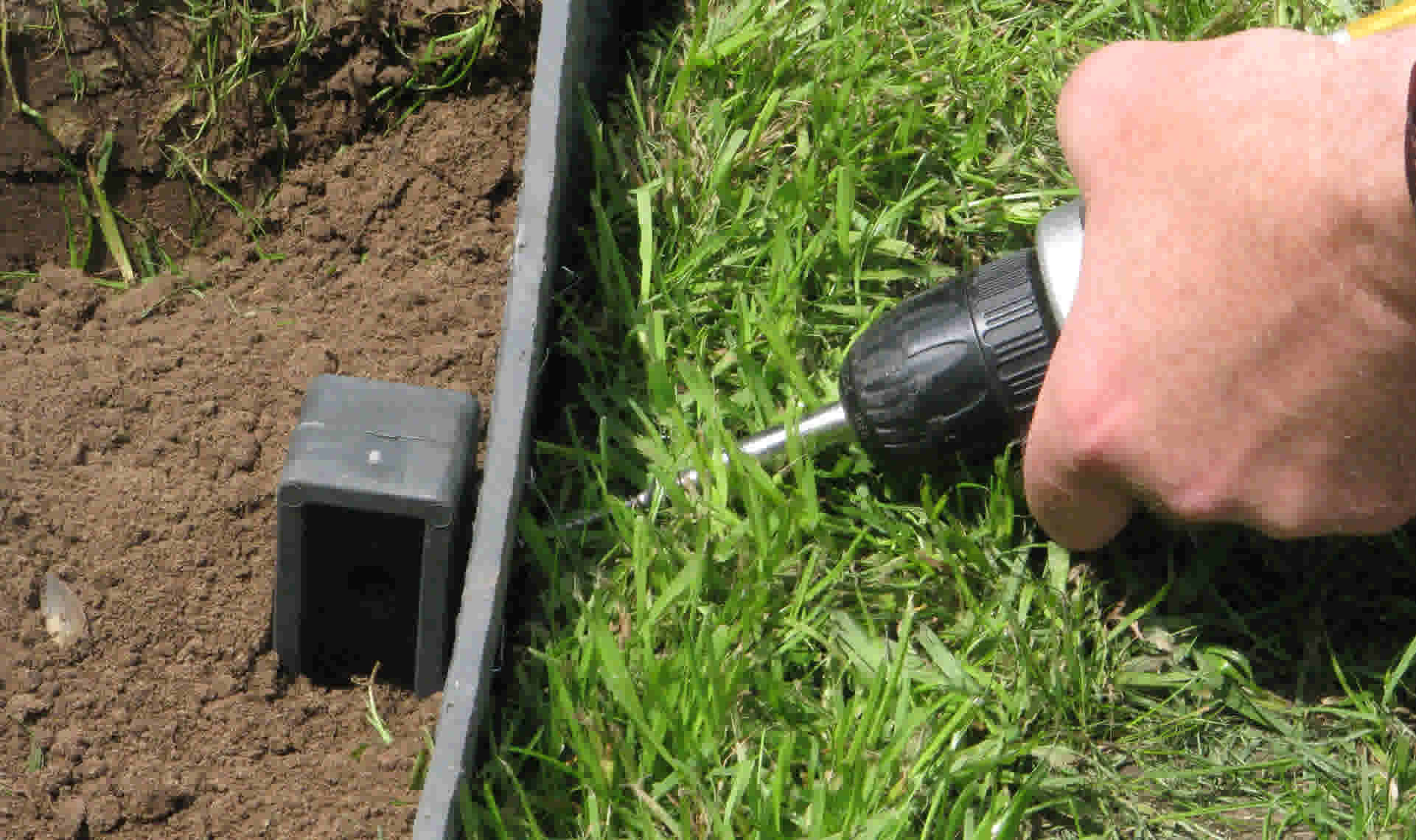 Hand gebruikt een boormachine om een tuinafboording te installeren tussen gras en aarde, met een zwart verbindingsstuk zichtbaar.