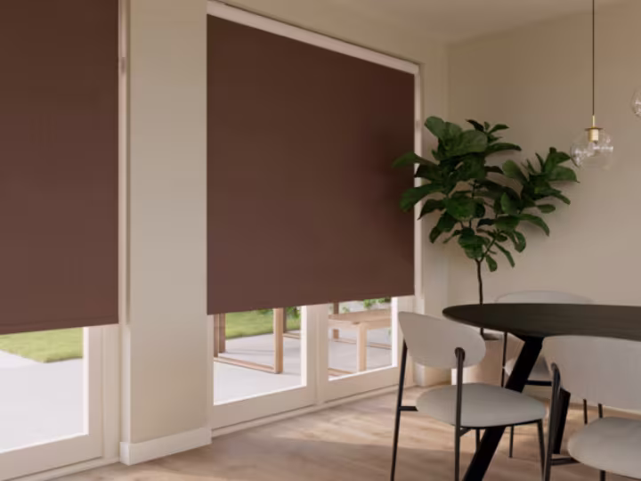Eetkamer met bruine rolgordijnen voor grote ramen, een ronde donkere tafel, stoelen en een groene plant in de hoek.