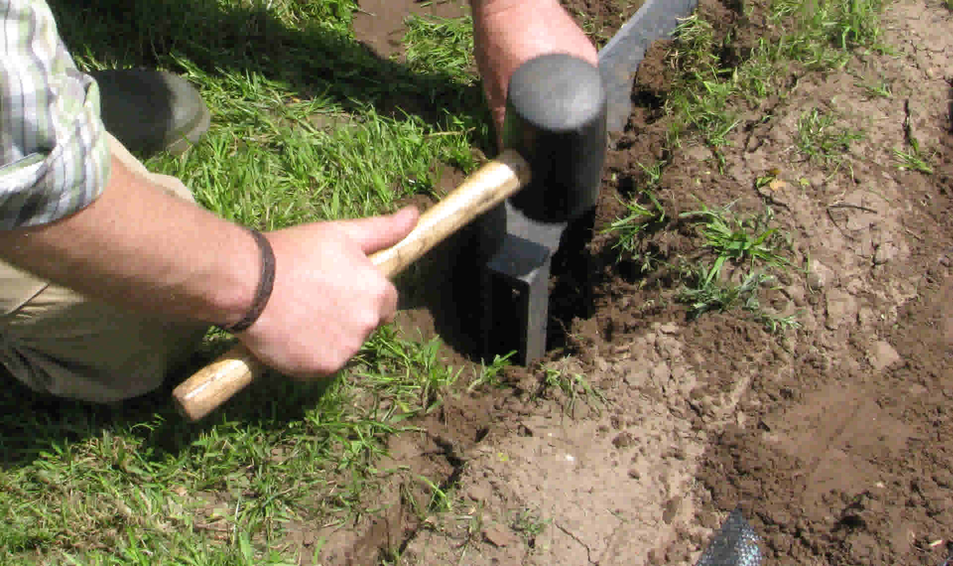 Persoon gebruikt een rubberen hamer om een metalen tuinrand in de grond te slaan tussen gras en aarde.