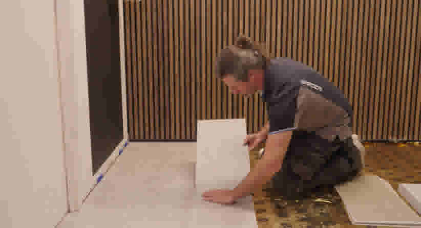 Vakman in werkkleding plaatst een clickvinyl op de vloer tegen een achtergrond van houten latten.