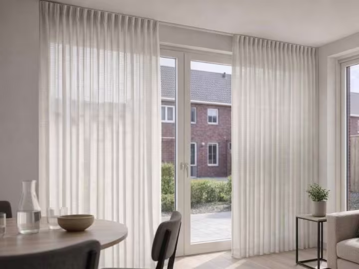 Lichte woonkamer met witte gordijnen voor glazen deuren die uitkijken op bakstenen huizen, met een eettafel en plantje op bijzettafel.