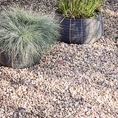 Deux pots en osier noir contenant des herbes ornementales vertes, posés sur un sol recouvert de gravier beige et gris.