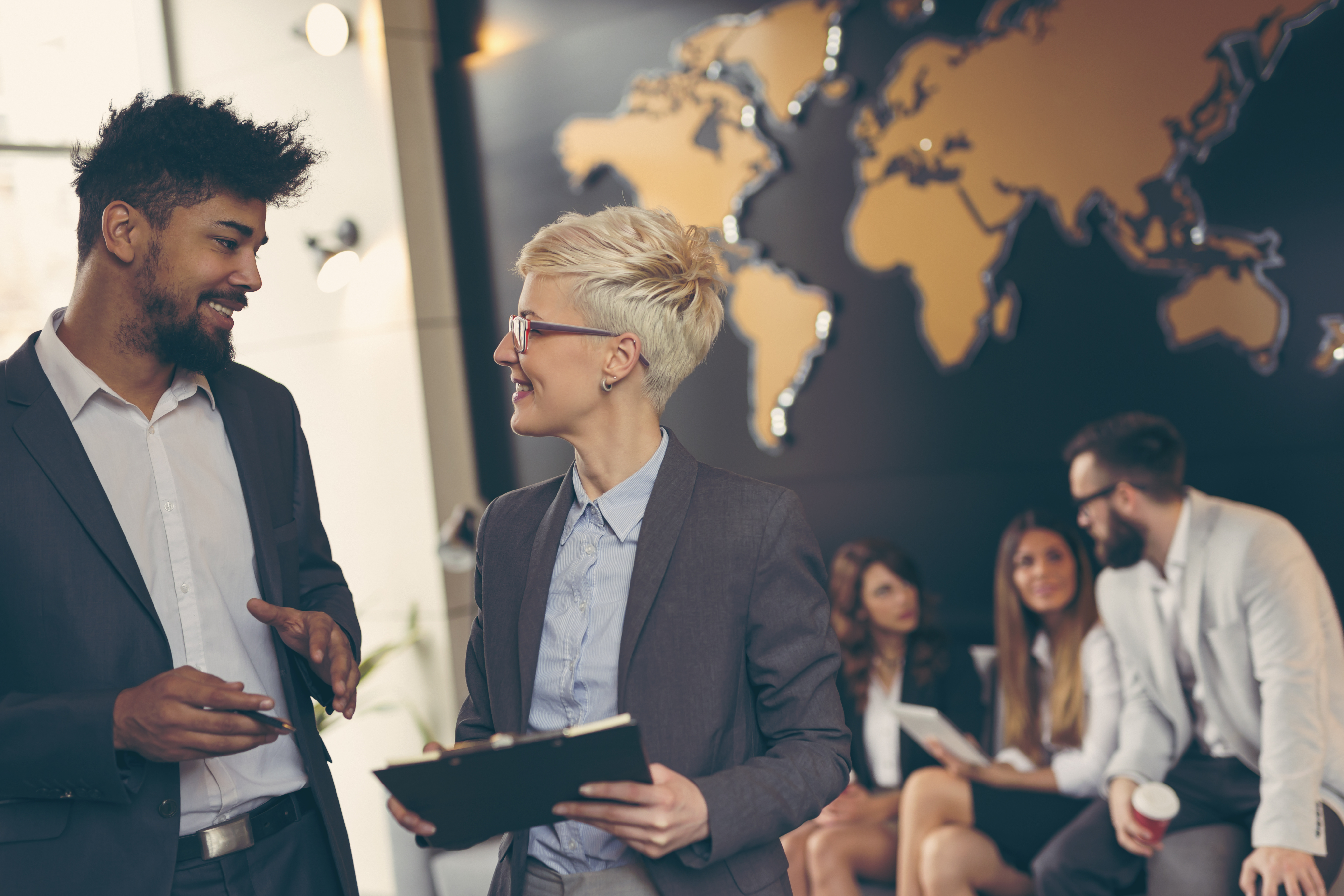 Two professional individuals have a discussion in front of some other working professionals