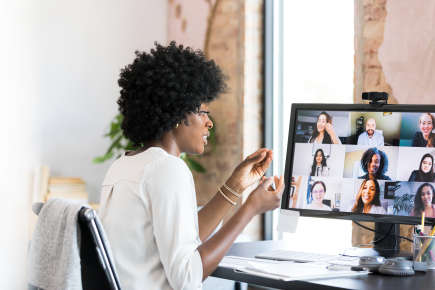 A mid adult businesswoman gestures while teleconferencing with colleagues. She is using her desktop PC in her home office.
