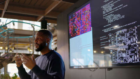 Man gives presentation with screens behind him displaying coding