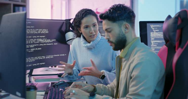 Computer programmer explaining code on a computer to a trainee programmer.