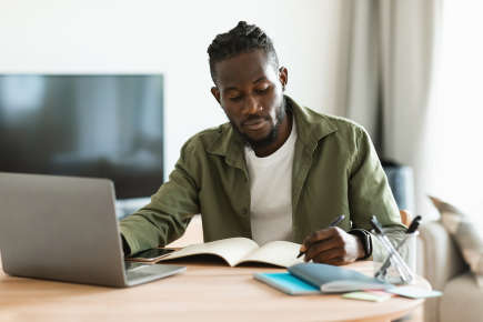 Focused african american man sitting at desk working on laptop and taking notes in notebook, studying online at home. Male watching webinar and using computer