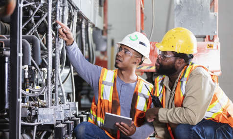 Two mid adult multiracial men inspecting the electrical components of an industrial refrigeration system. They are wearing hardhats, safety glasses and safety vests. 