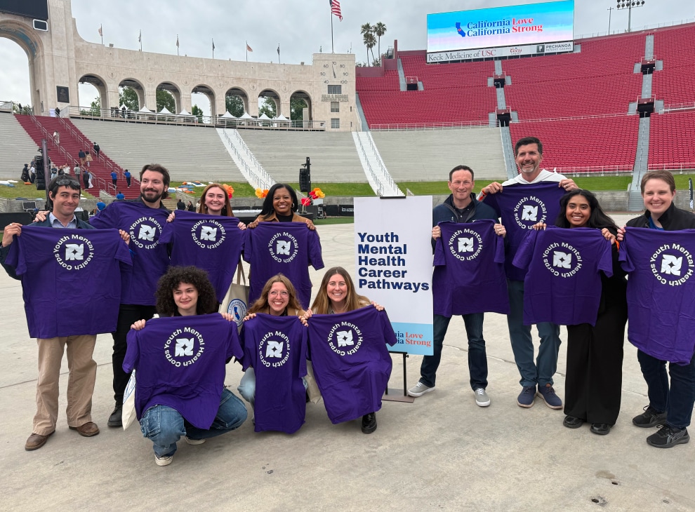 Group of adults smiling and holding up purple “Youth Mental Health Corps” t-shirts while posing in a stadium.