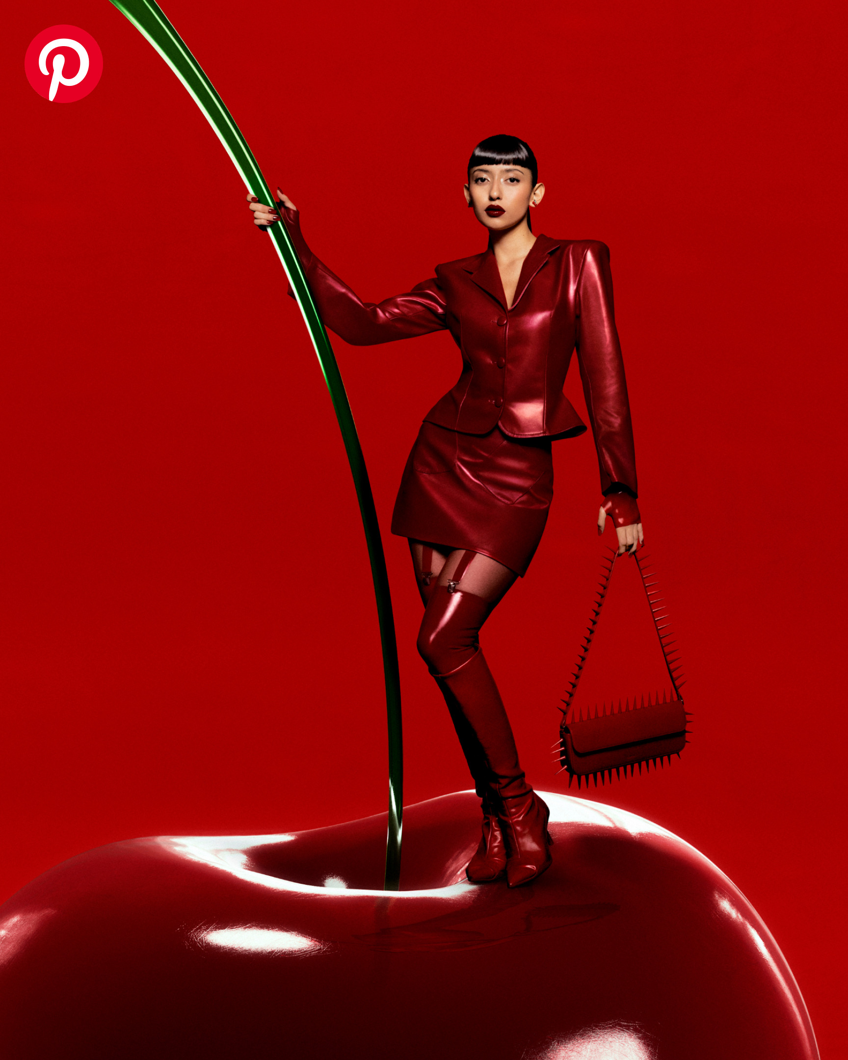 A woman with sleek black hair, dark red lipstick and a head-to-toe red leather outfit poses on top of a giant cherry