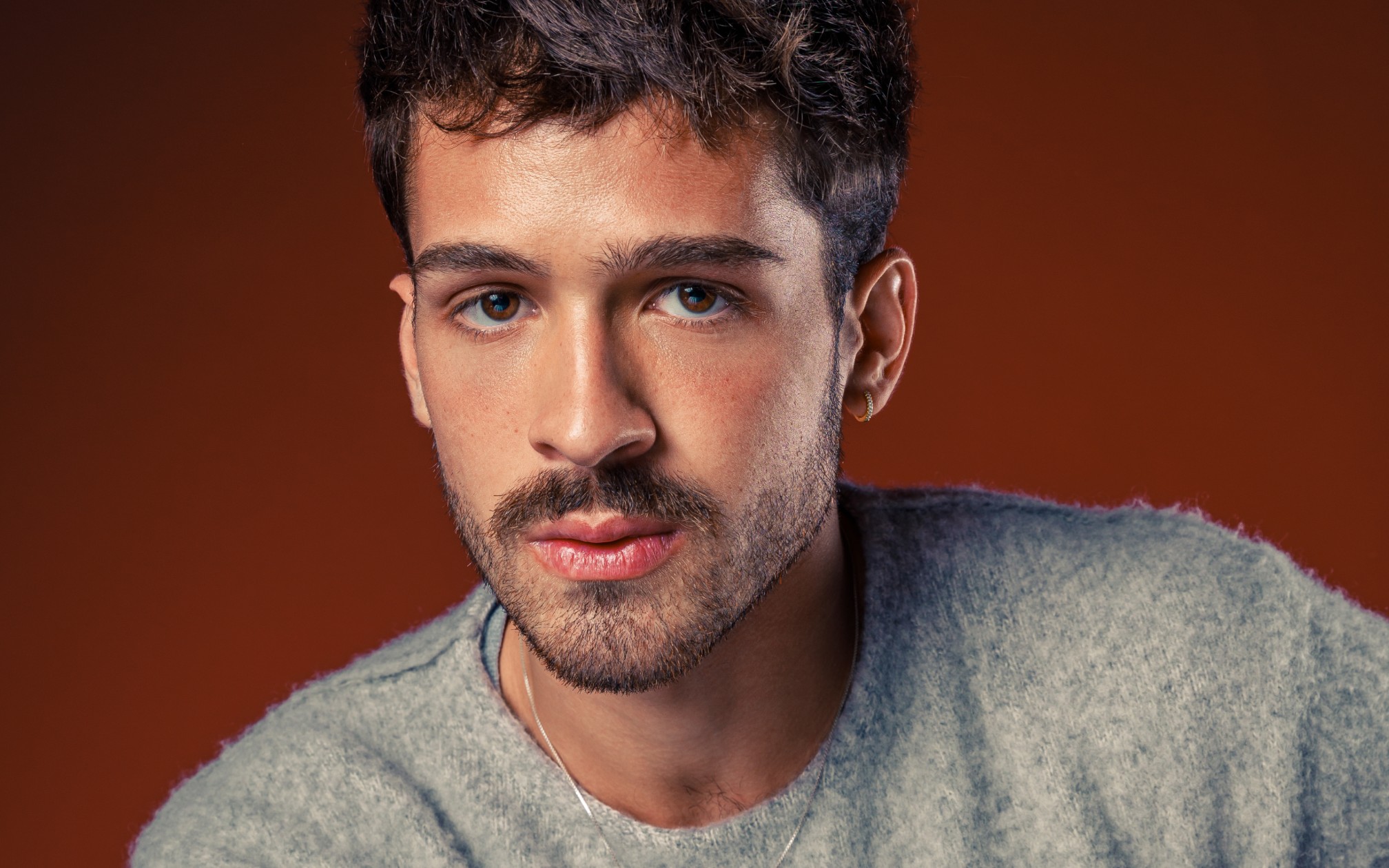Na imagem, João Guilherme vestindo um suéter cinza, com um colar, e fundo marrom